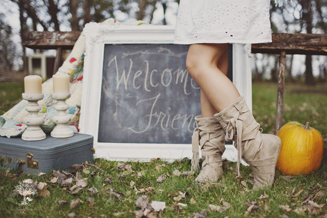 Chalkboard sign with "Welcome Friend" in white ornate frame.  Vintage suit case with 2 candle sticks and a pumpkin on the grass with girl's legs wearing boots