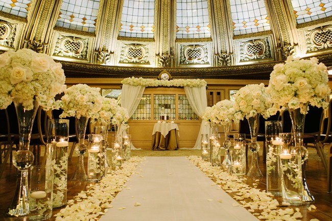 Ceremony for Arctic Club Hotel wedding: aisle with vases of white roses and floating candle, white aisle runner line with white rose pedals 