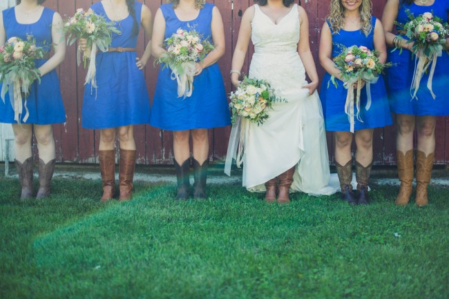 Bride standing with bridesmaids in blue dresses with brown skinny belt all wearing brown colored cowboy boots on grass