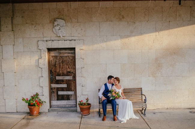 bride and groom sitting on a bench beside a wooden door and potted plants