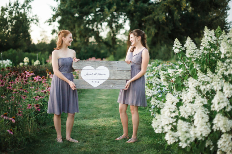 two bridesmaids in lavender dresses hold a wood sign